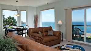 A living room with a brown leather sofa, a dining table with chairs, a lamp, and a view of the ocean through large windows. - Keystone Vacation Rentals-Ocean View Condos (Depoe Bay)