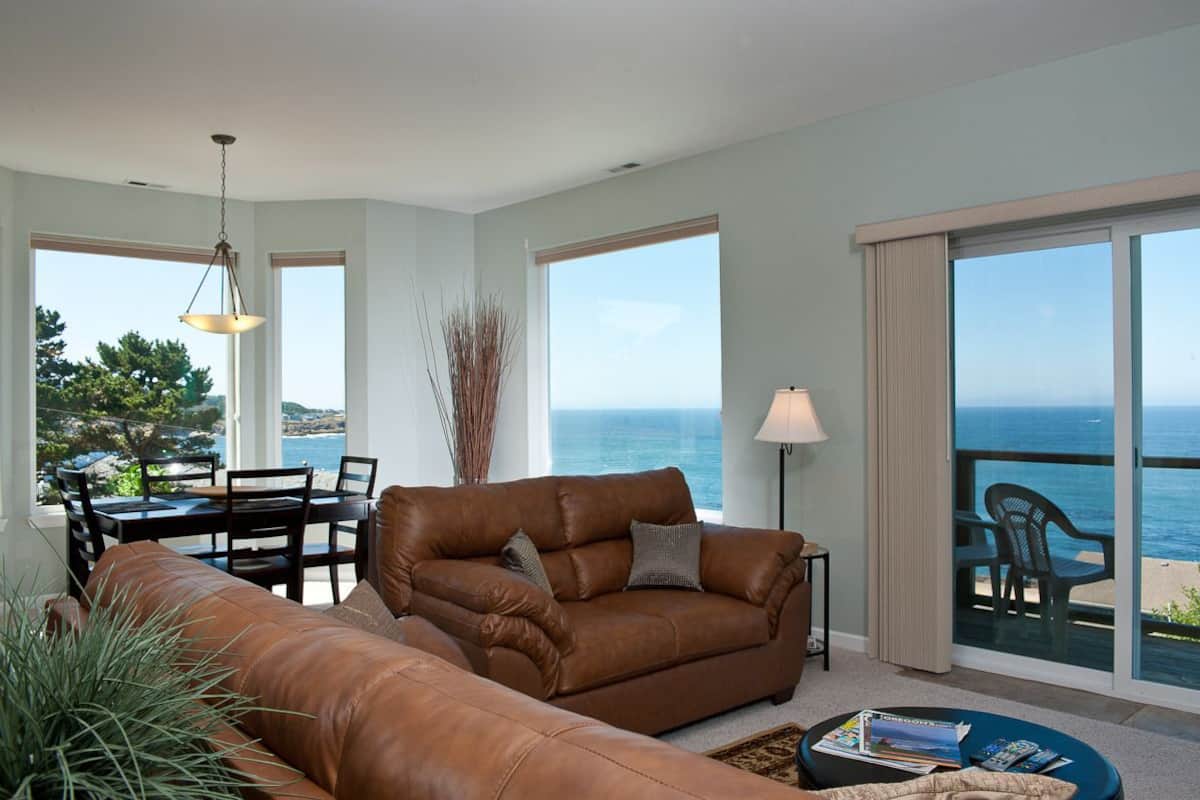 A living room with a brown leather sofa, a dining table with chairs, a lamp, and a view of the ocean through large windows.