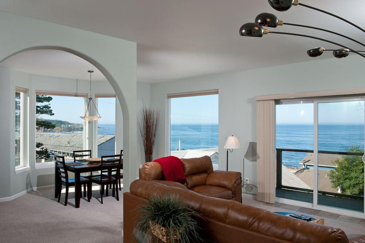 A spacious living room with a large window overlooking the ocean, a dining area with a table and chairs, and a cozy seating area with a leather sofa.
