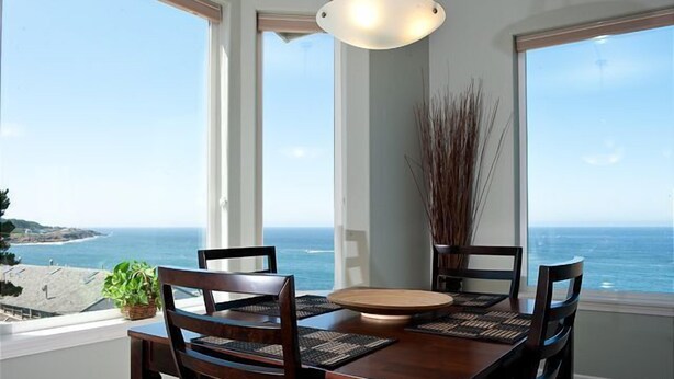 A dining area with a view of the ocean, featuring a wooden table and chairs, a hanging light fixture, and a potted plant. - Keystone Vacation Rentals-Ocean View Condos (Depoe Bay)