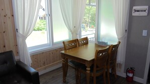 A dining area with a wooden table and chairs, large windows with white curtains, a fire extinguisher, and a thermostat on the wall.