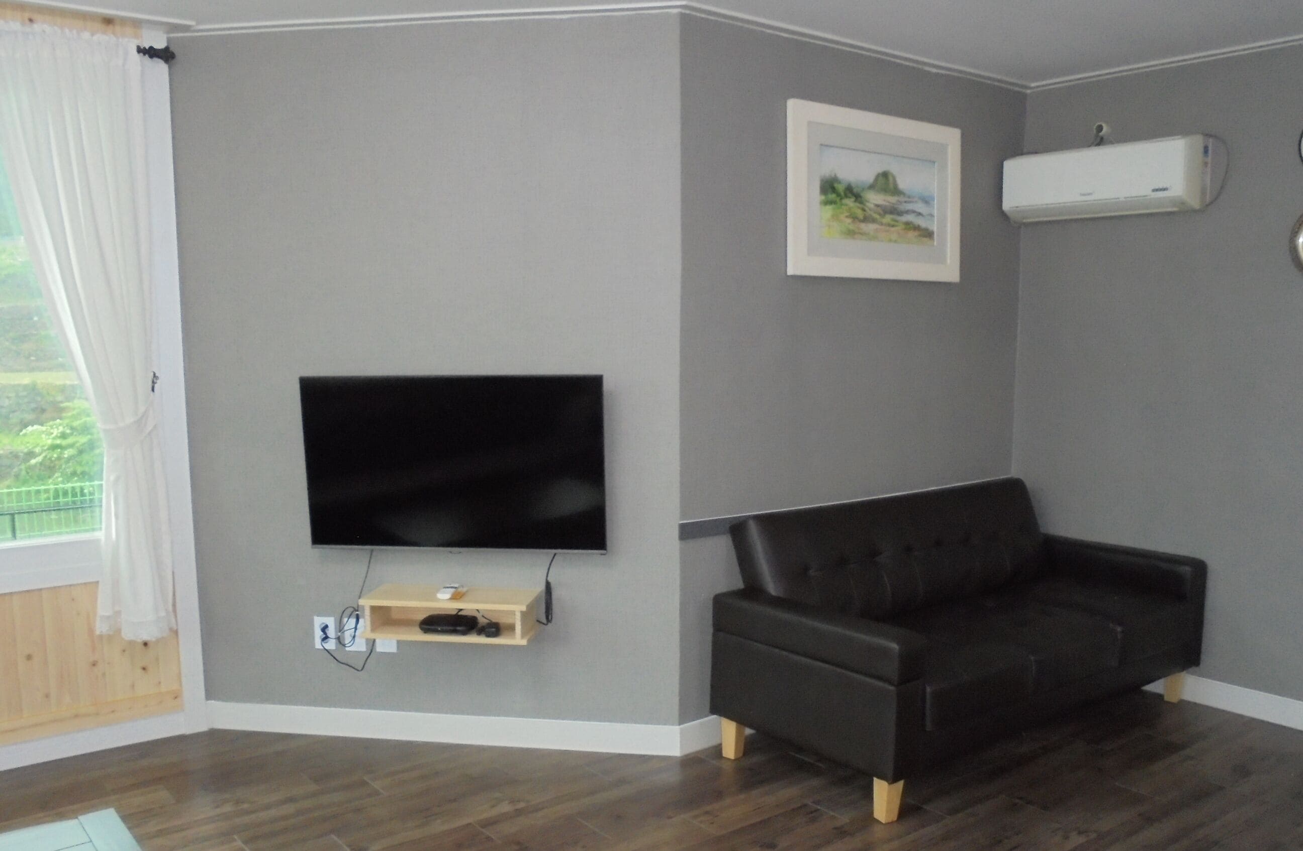 A living room with a flat-screen TV mounted on the wall, a black leather sofa, and a small wooden shelf with electronic devices.