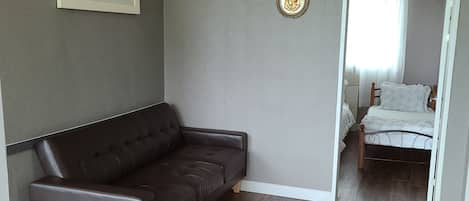 A living room with a dark brown leather sofa, a wall-mounted air conditioner, a clock, and a door leading to a bedroom with a bed and a nightstand.