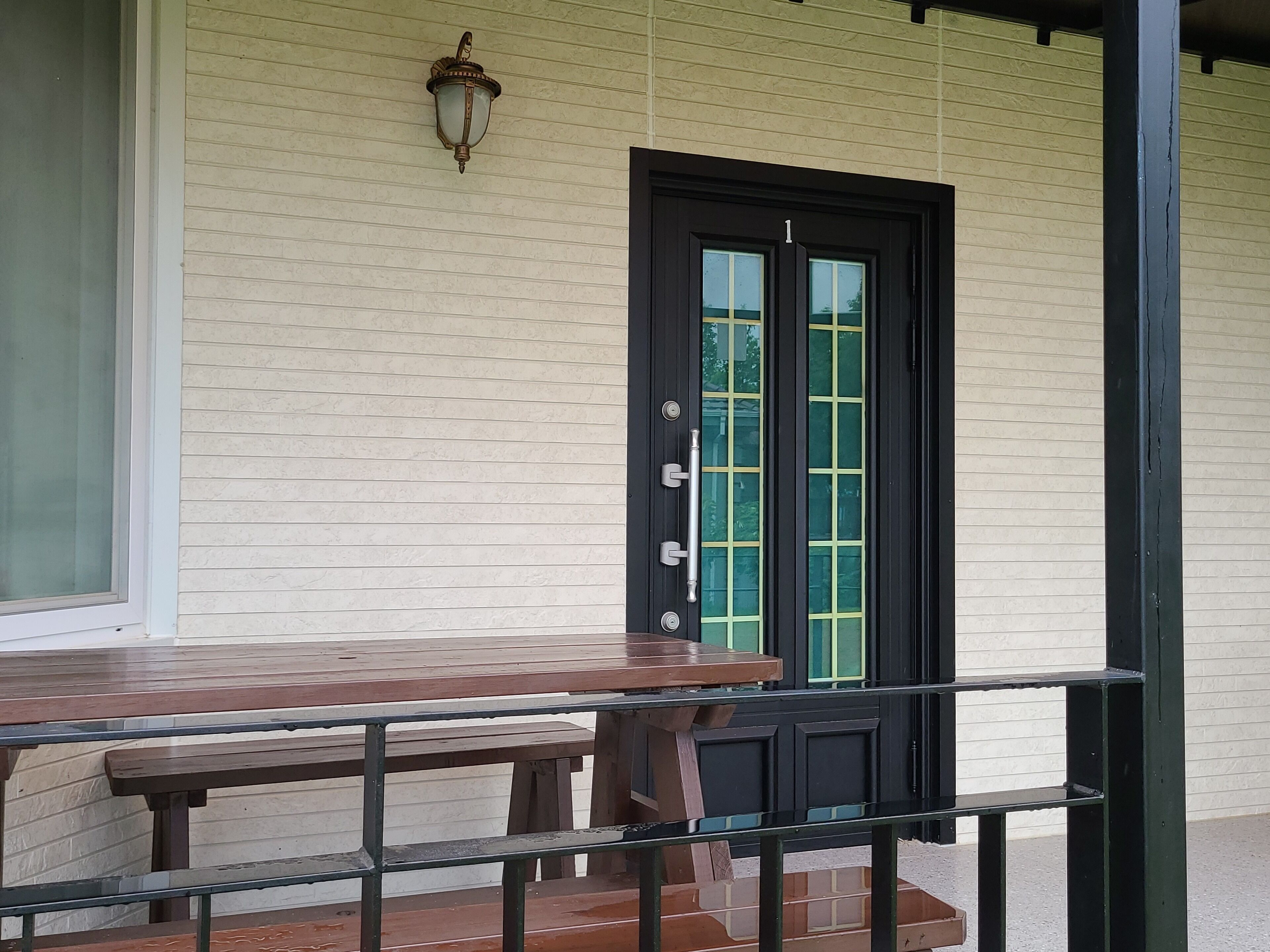 A wooden table and bench on a porch with a black door and a light fixture above it.