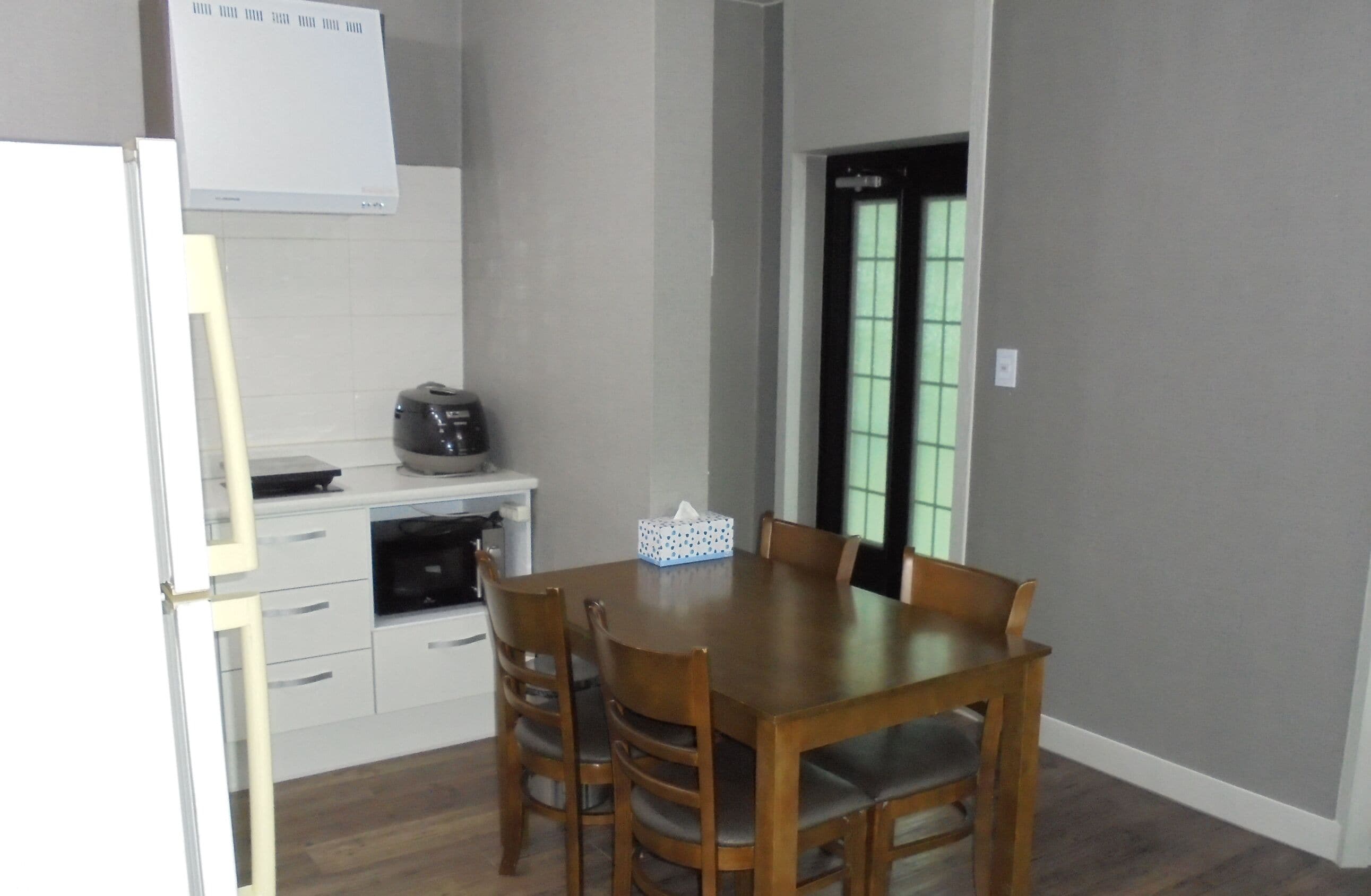 Un comedor con una mesa y sillas de madera, una pequeña cocina con refrigerador y microondas, y un ventanal con vidrio tintado de verde.