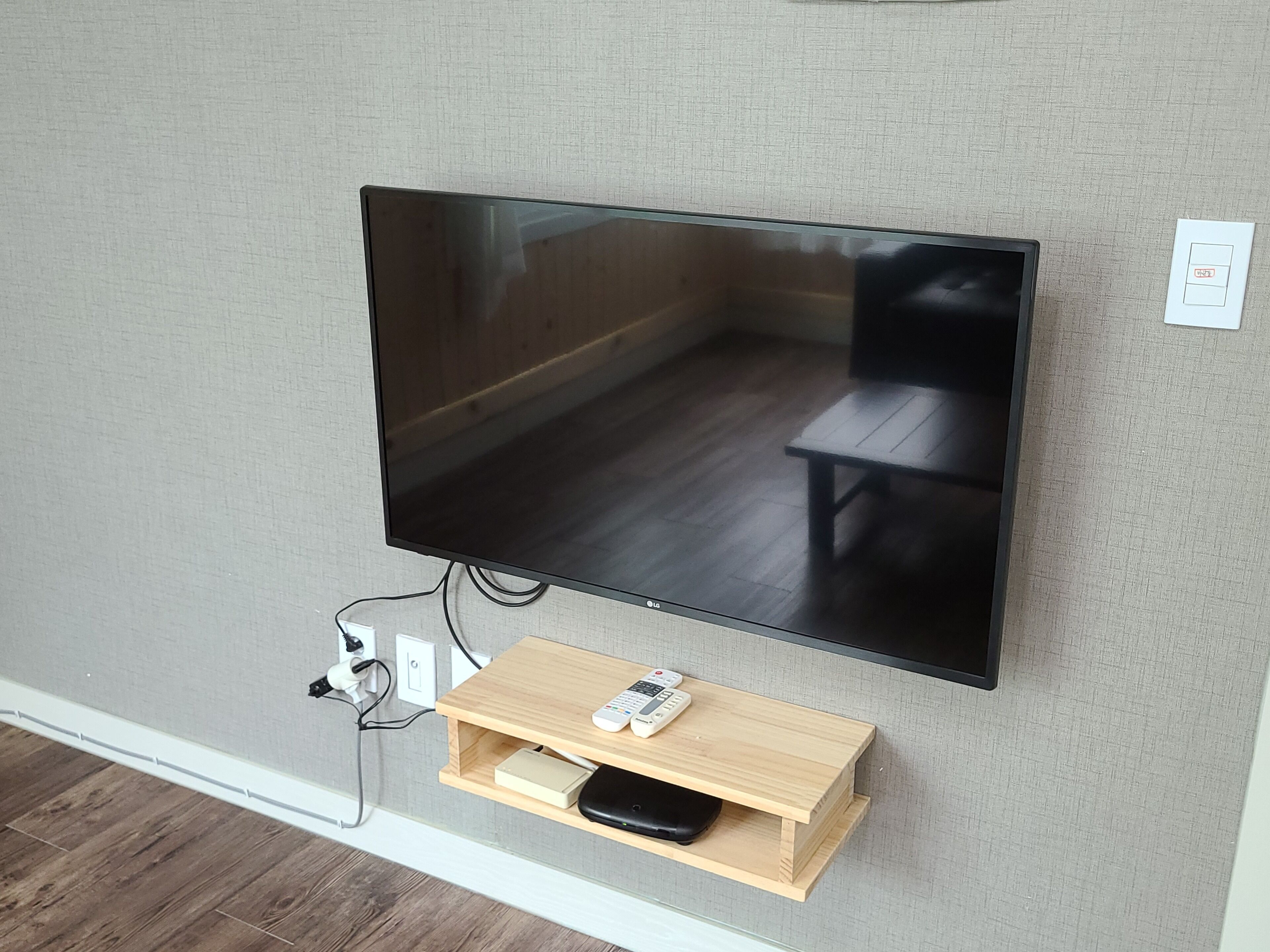 A flat-screen TV mounted on a textured wall, connected to a wooden shelf below holding a remote and a black device.