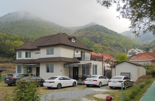 A two-story house with a garage, parked cars, and a mountainous backdrop. - Santa Fe Pension (Namhae)