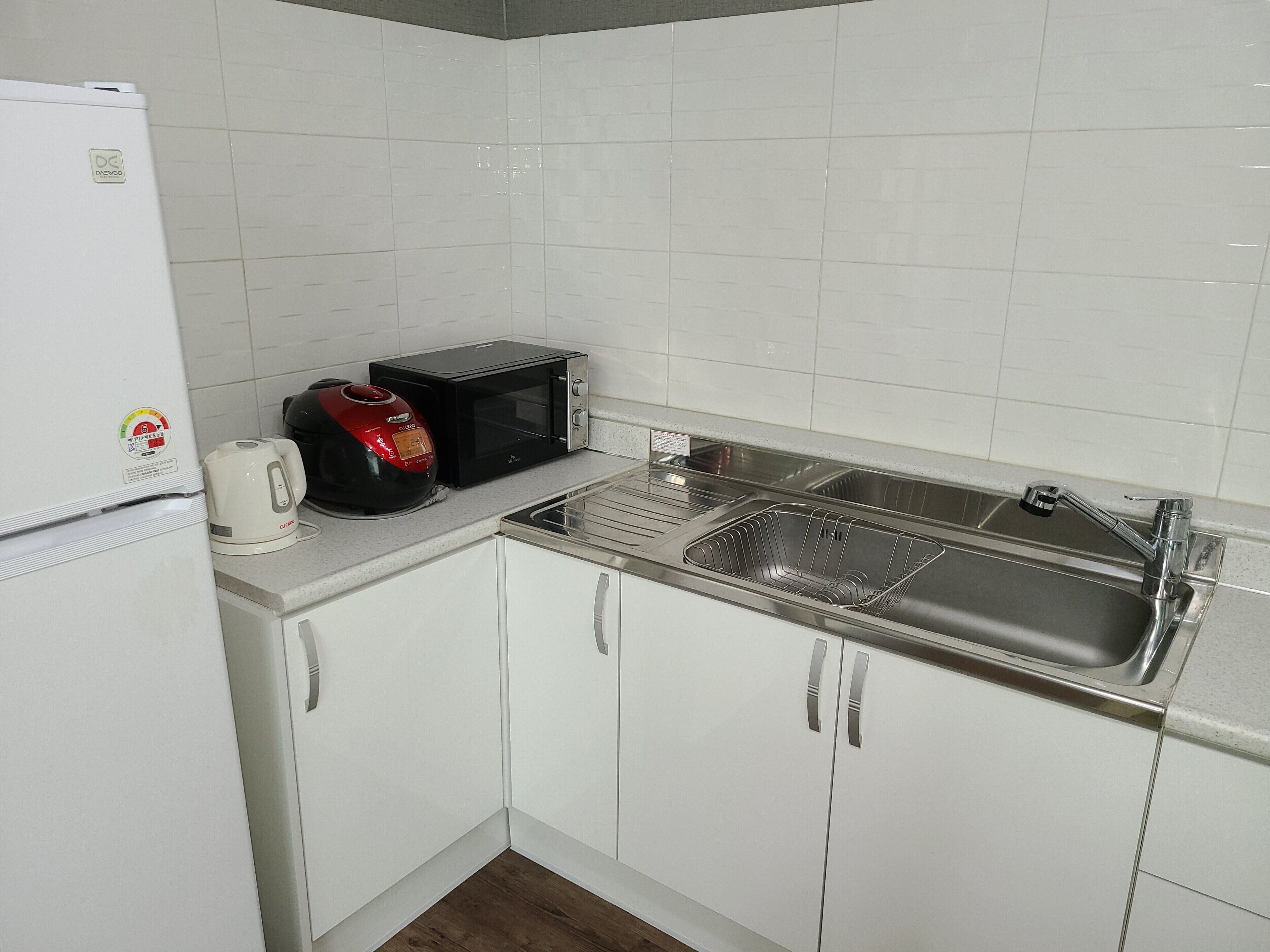 Una pequeña cocina con armarios blancos, un lavamanos de acero inoxidable, un microondas y una tetera.