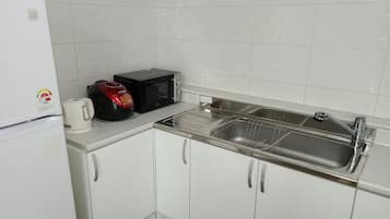 Una pequeña cocina con armarios blancos, un lavamanos de acero inoxidable, un microondas y una tetera.