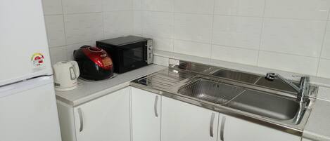 Una pequeña cocina con armarios blancos, un lavamanos de acero inoxidable, un microondas y una tetera.