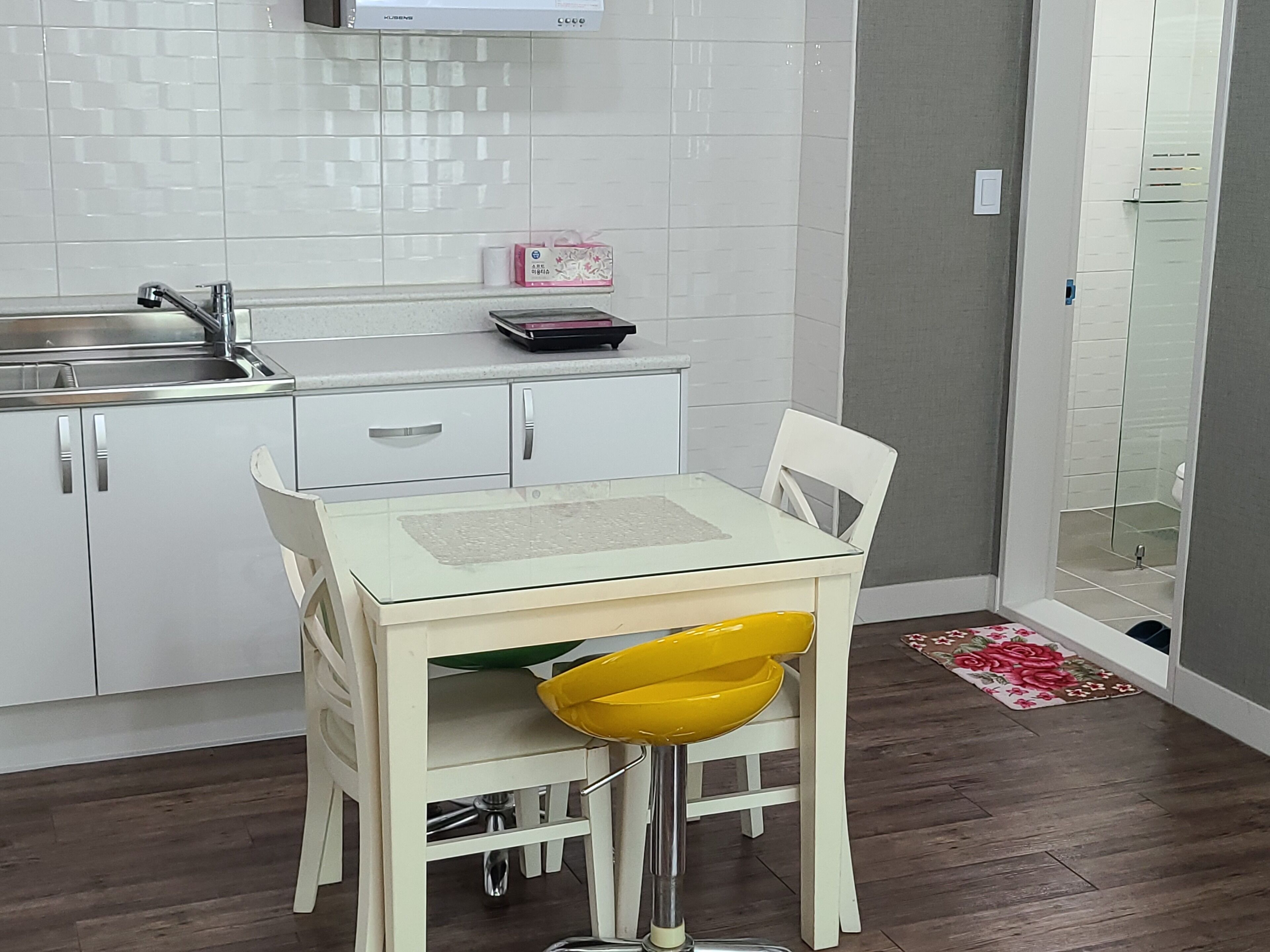 A compact kitchen with white cabinets, a stainless steel sink, and a small countertop. A white table with a glass top and a yellow chair is positioned in front.