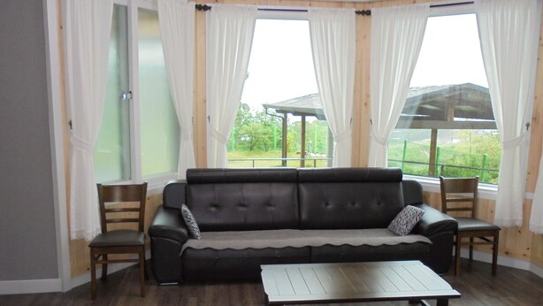 A room with a dark leather sofa, a wooden coffee table, and two wooden chairs. There are large windows with white curtains, and a view of greenery outside.
