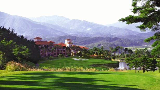 A golf course with a clubhouse, a pond, and mountains in the background. - Pinehills Hotel (Suncheon)