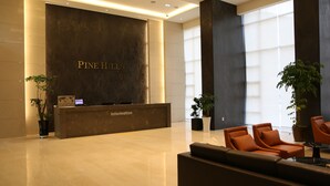 A reception area with a dark reception desk labeled "Pine Hills Information," two potted plants, and a seating area with orange chairs. - Pinehills Hotel (Suncheon)