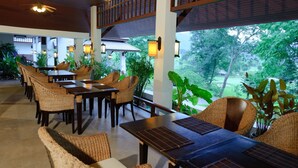 An indoor dining area with wicker chairs and tables, large windows with a view of greenery, and ceiling fans. - Chiangmai Highlands Golf and Spa Resort (Mae On)