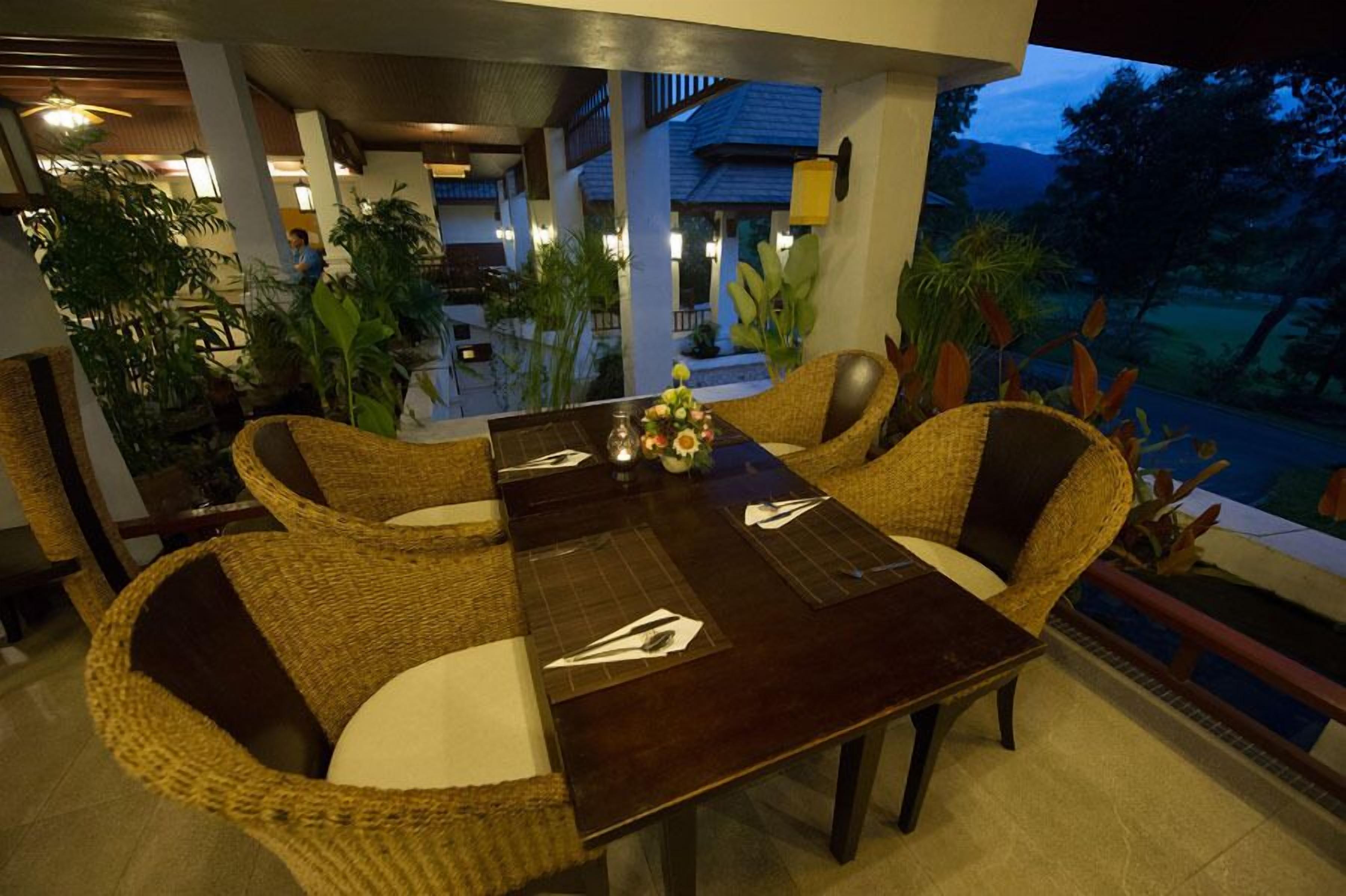 Un espacio de comedor al aire libre con sillas de mimbre y una mesa de madera oscura, rodeado de plantas en macetas y con vistas a un paisaje pintoresco.