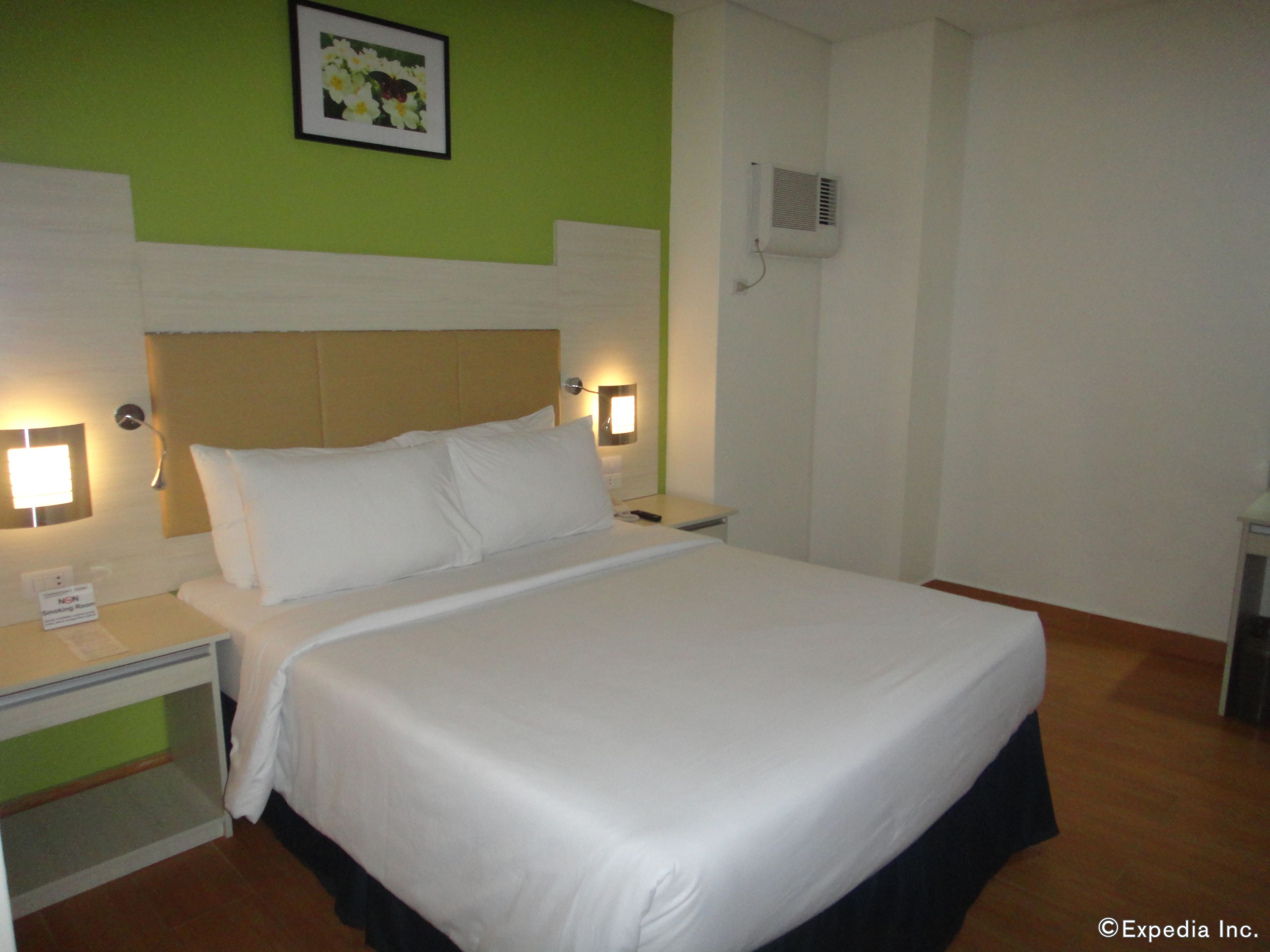 A hotel room with a large bed, two bedside tables, a nightstand, a wall-mounted air conditioner, and a framed picture on the wall.