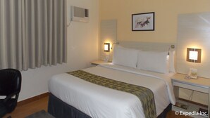 Una habitación de hotel con cama, una silla, una mesita de noche con teléfono y aire acondicionado instalado en la pared.