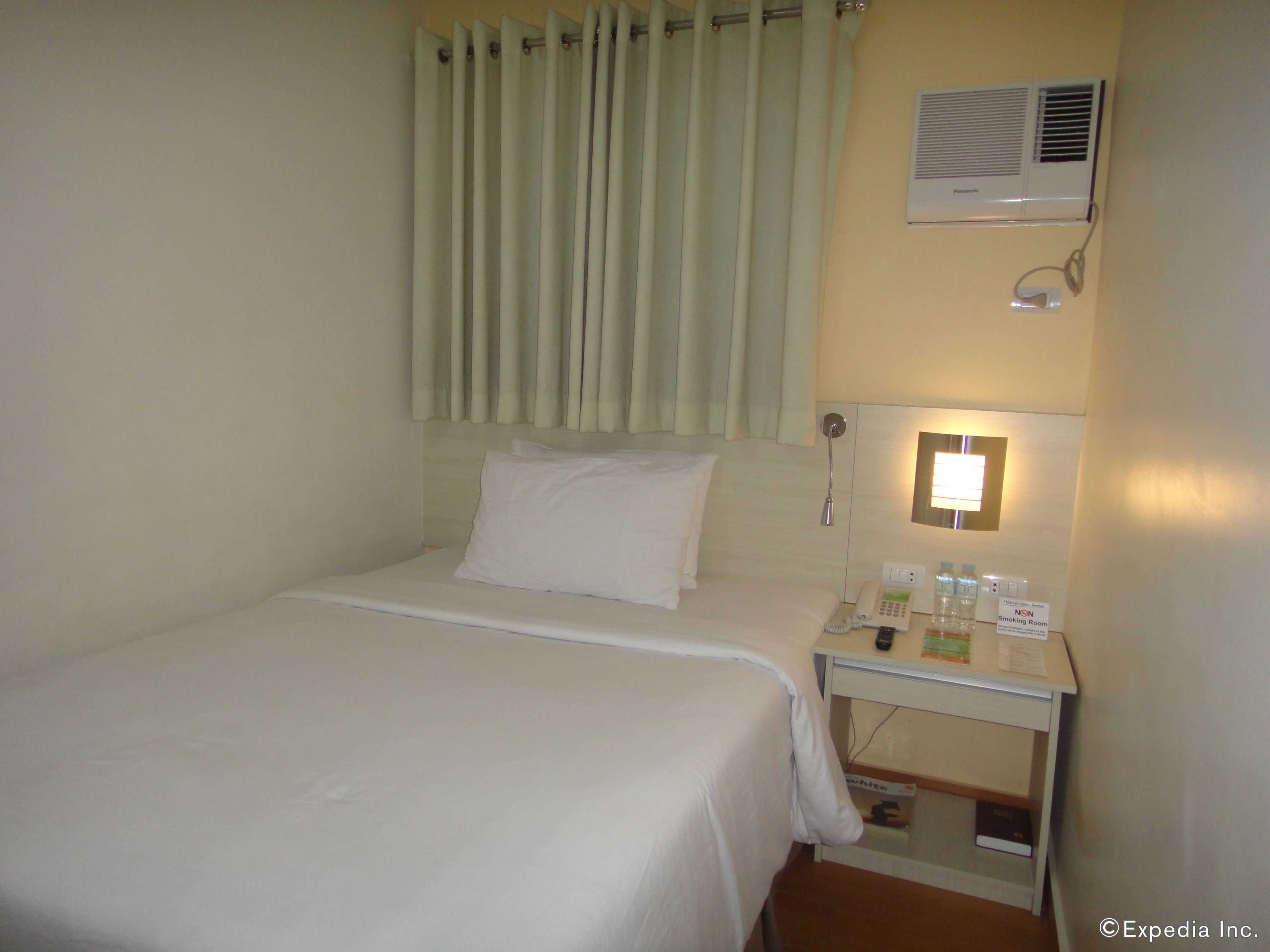 Habitación de hotel con cama, mesita de noche con teléfono y una botella, cortina, equipo de aire acondicionado y un letrero que indica que es para fumadores.