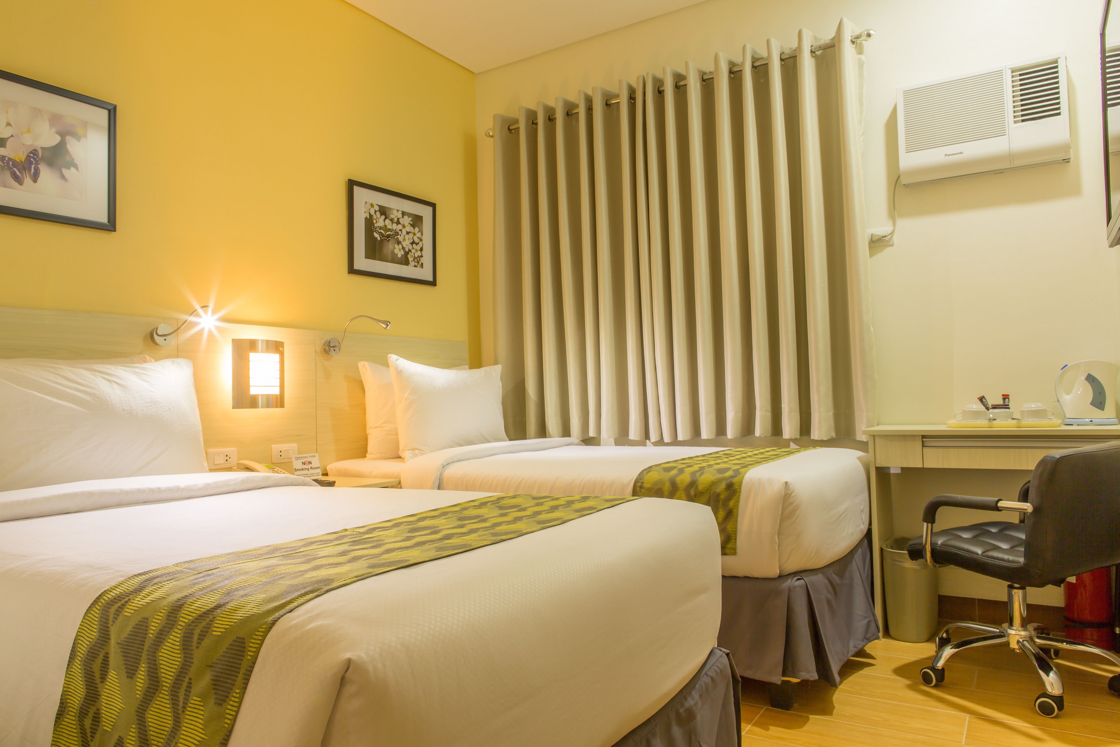 Habitación de hotel con dos camas, un escritorio, una silla, un cubo de basura y una ventana con cortinas.