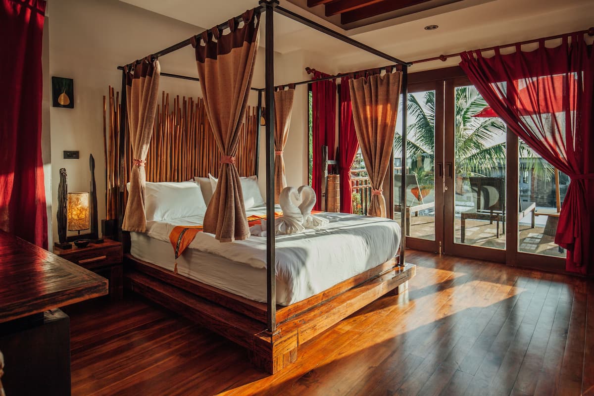 Una cama con dosel y cortinas, suelo de madera y vista a palmeras a través del ventanal.