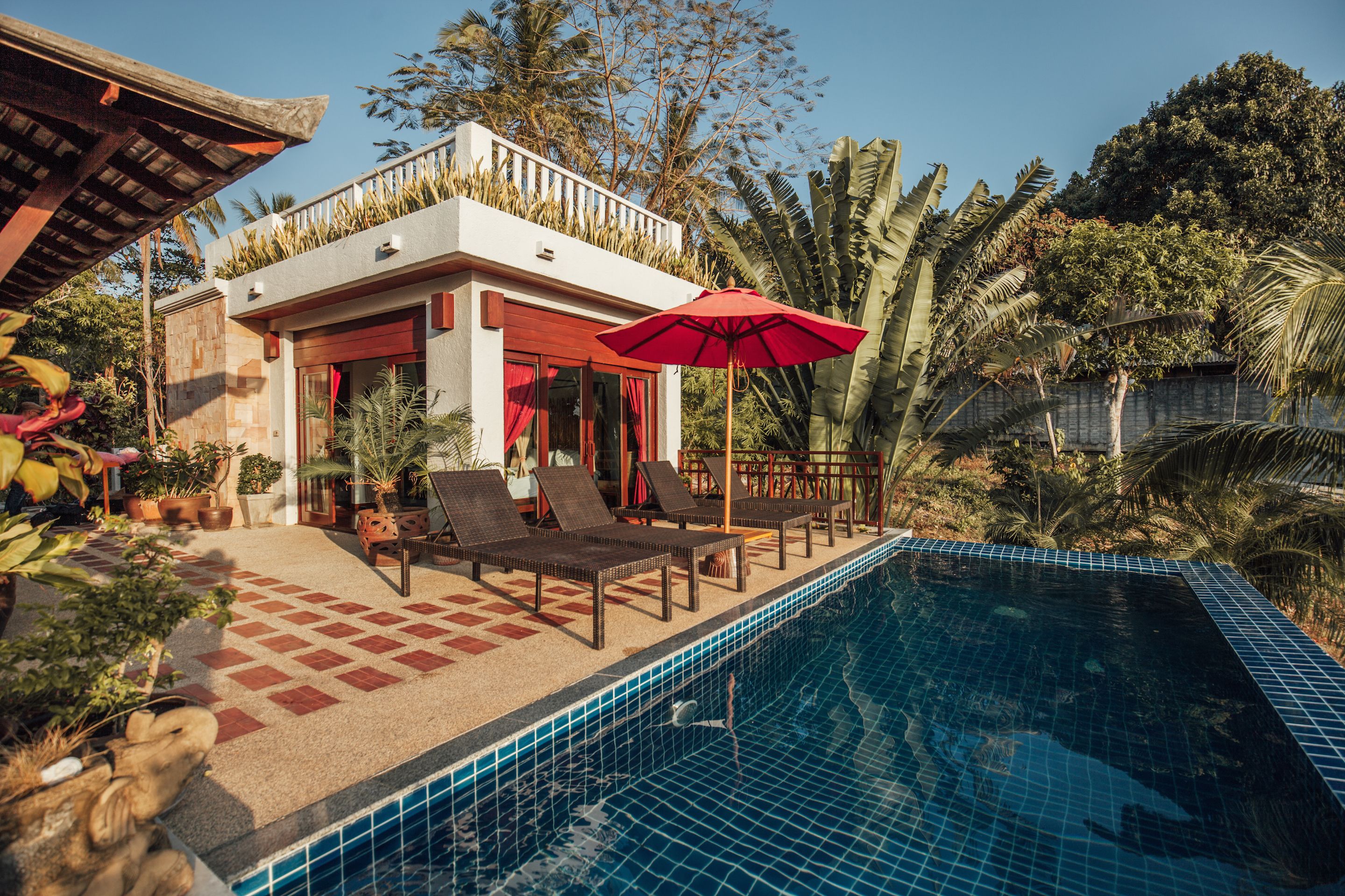 A poolside area with lounge chairs and a red umbrella, surrounded by tropical plants and a clear blue pool.
