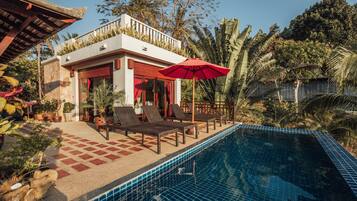 A poolside area with lounge chairs and a red umbrella, surrounded by tropical plants and a clear blue pool.