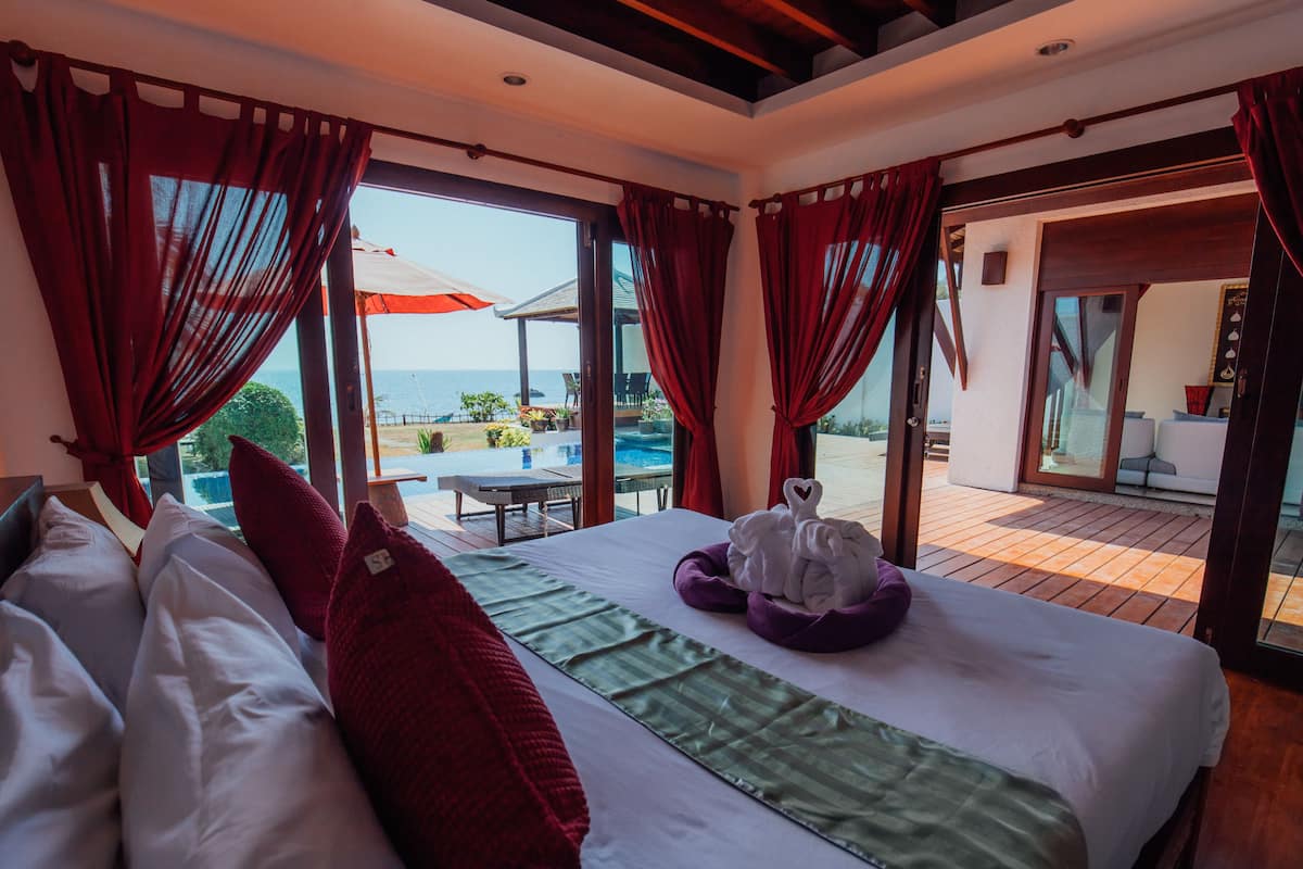 Un dormitorio con cama, cortinas rojas y vista a la piscina y zona de estar exterior.