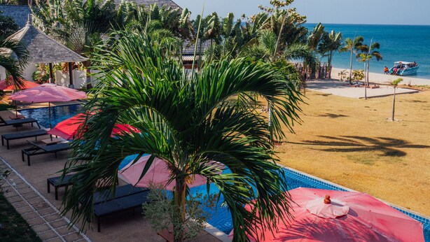 A pool area with lounge chairs and umbrellas, a beach with people, and a boat on the water. - Thai Island Dream Estate (Ko Lanta)
