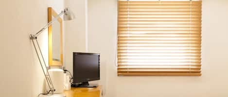 A hotel room with a desk, a computer, a bed, a window with blinds, an air conditioner, and a green trash bin.