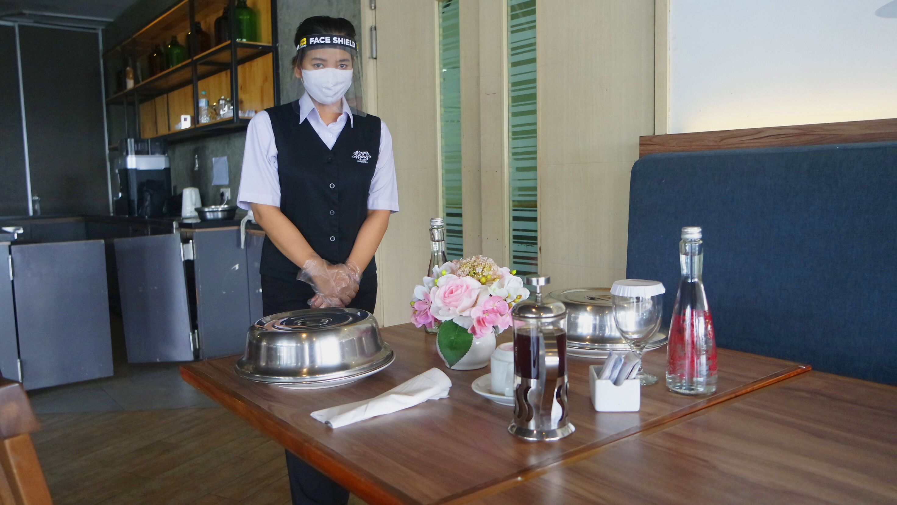 Un miembro del personal de un hotel preparando una mesa con una bandeja plateada, un jarrón de flores y diferentes vasos.