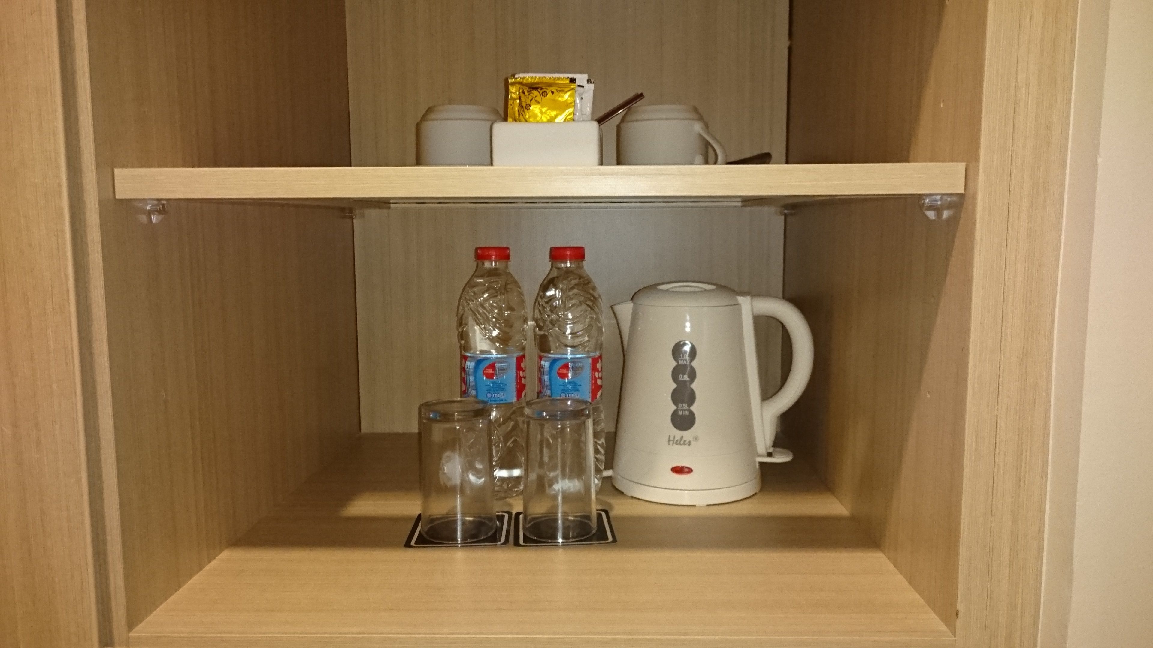 A wooden cabinet with a shelf containing two bottles, two glasses, and a white electric kettle.