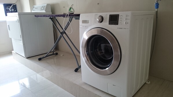 A front-loading washing machine with a digital display and control panel, a drying rack, and a smaller appliance in the background. - Ayaka Suites (Jakarta)