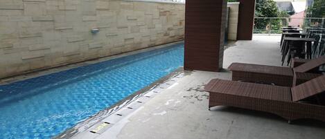 An indoor swimming pool with clear blue water, surrounded by a tiled floor and a stone wall. There are wicker lounge chairs and a table near the pool.