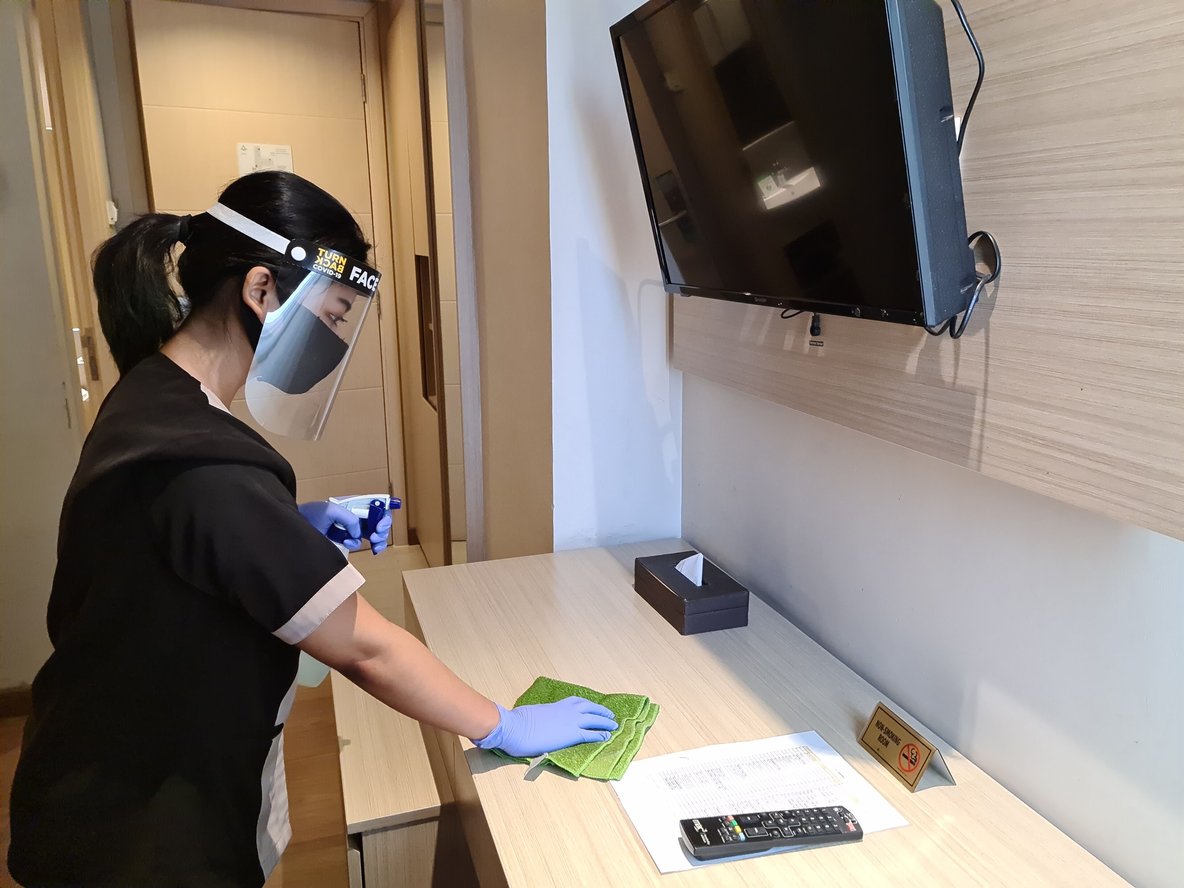 A person cleaning a wooden desk with a green cloth, a remote control, and a tissue box.