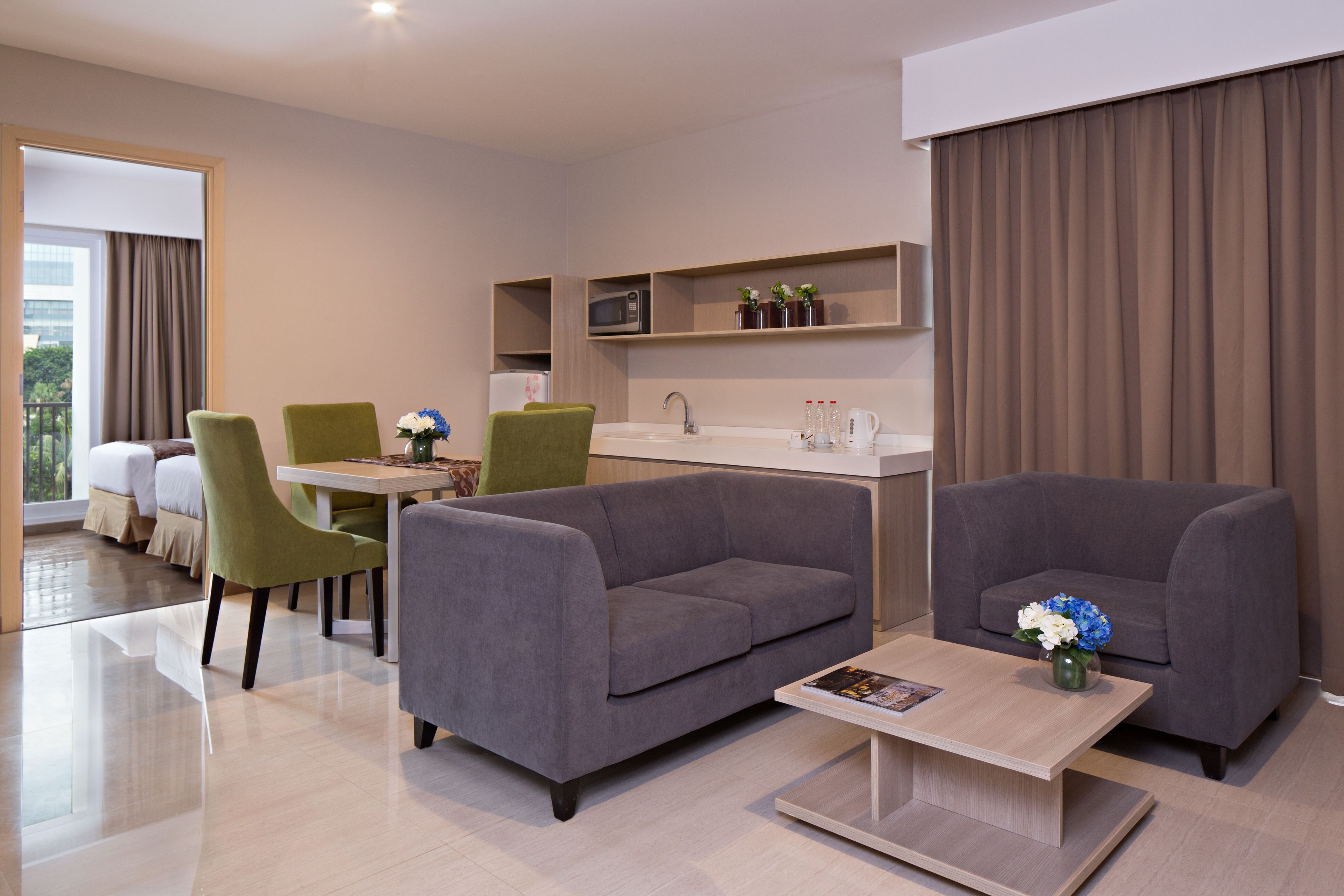 Une chambre d’hôtel moderne avec un canapé gris, des fauteuils verts, une table à manger et un coin cuisine équipé d’un four à micro-ondes et d’un évier.