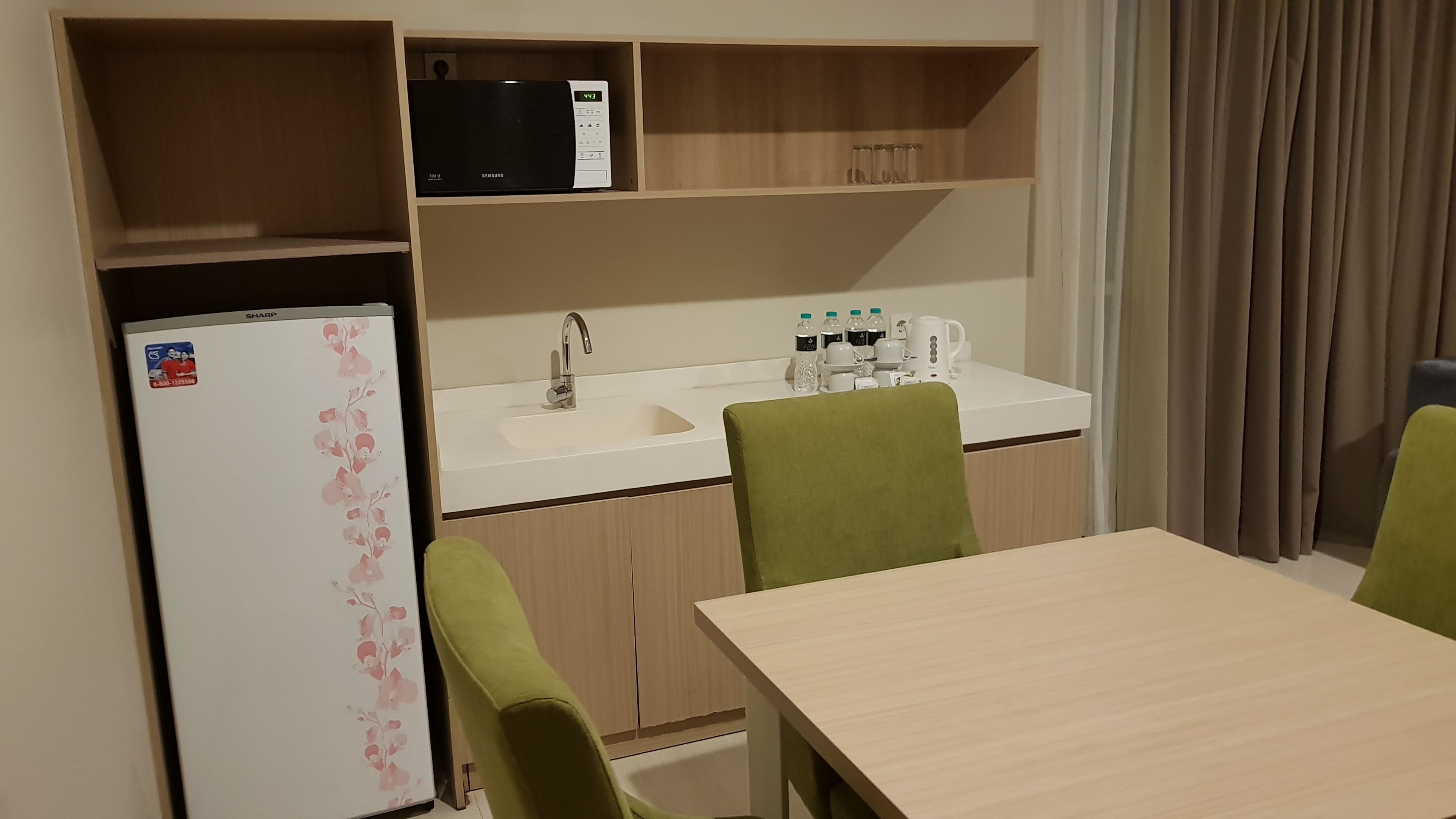 Een kleine kitchenette met een koelkast, magnetron en gootsteen. Er is een tafel met twee stoelen en een raam met gordijnen.