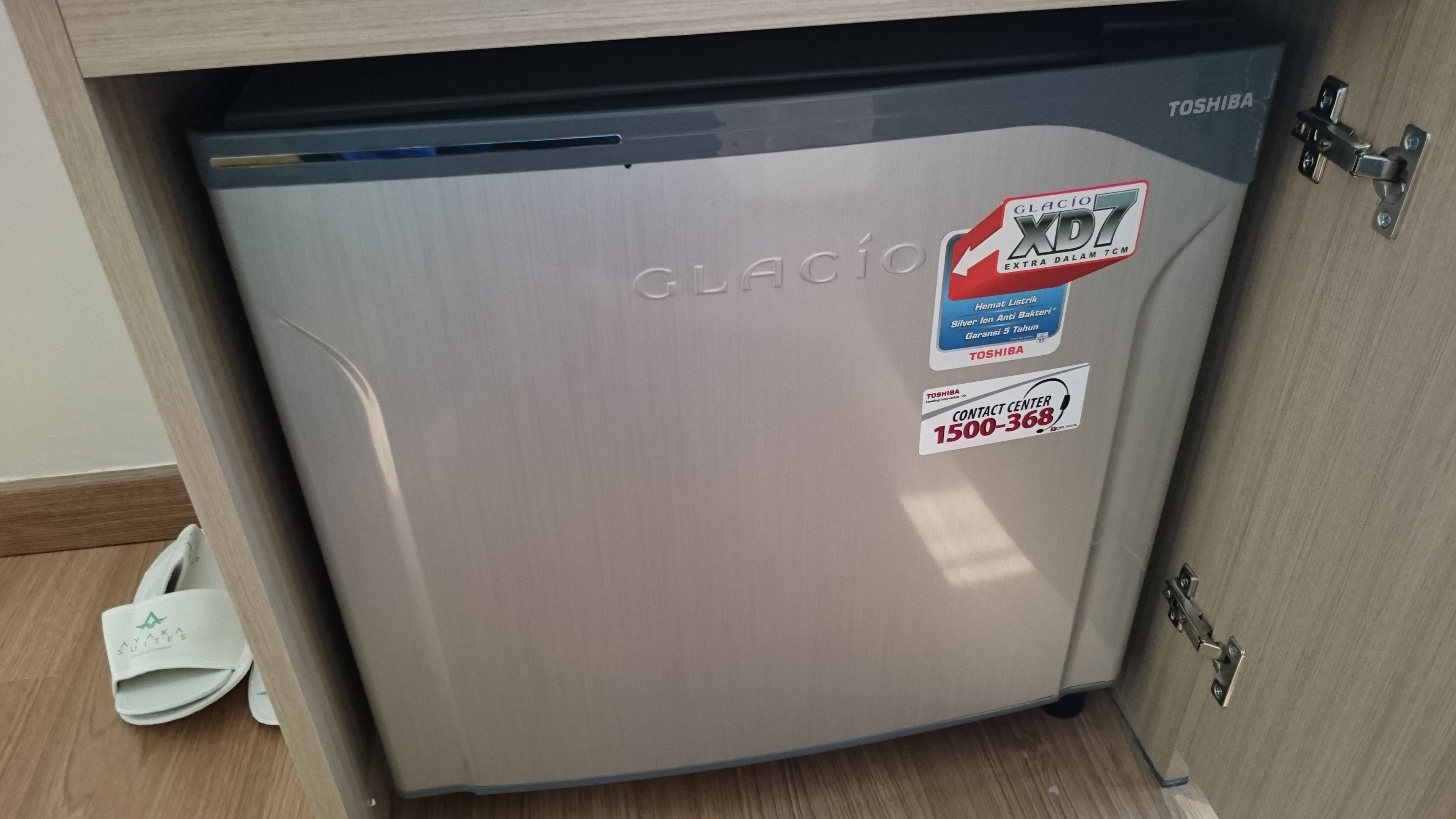 A compact, stainless steel refrigerator with a Toshiba logo, under a wooden cabinet.