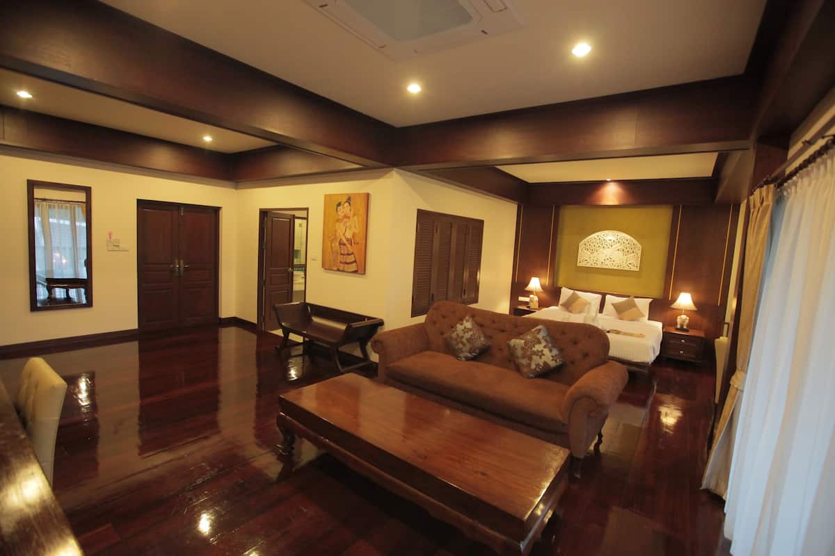A spacious living room with a large wooden coffee table, a brown sofa with patterned cushions, and a white armchair. There is a wooden cabinet and a painting on the wall.