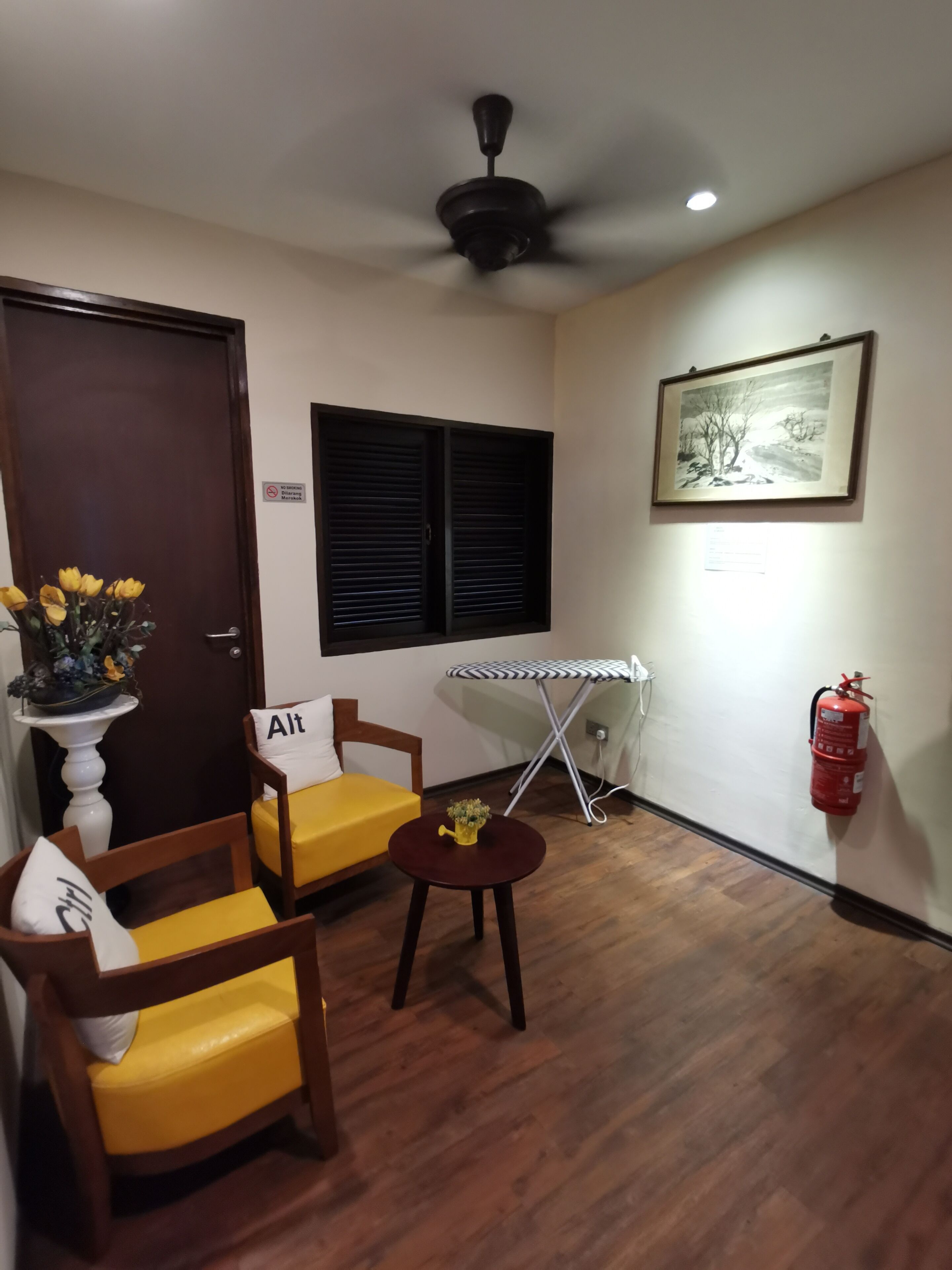A room with a wooden floor, a ceiling fan, a door with a sign, two yellow chairs, a small table with a plant, a framed picture on the wall, and a fire extinguisher.