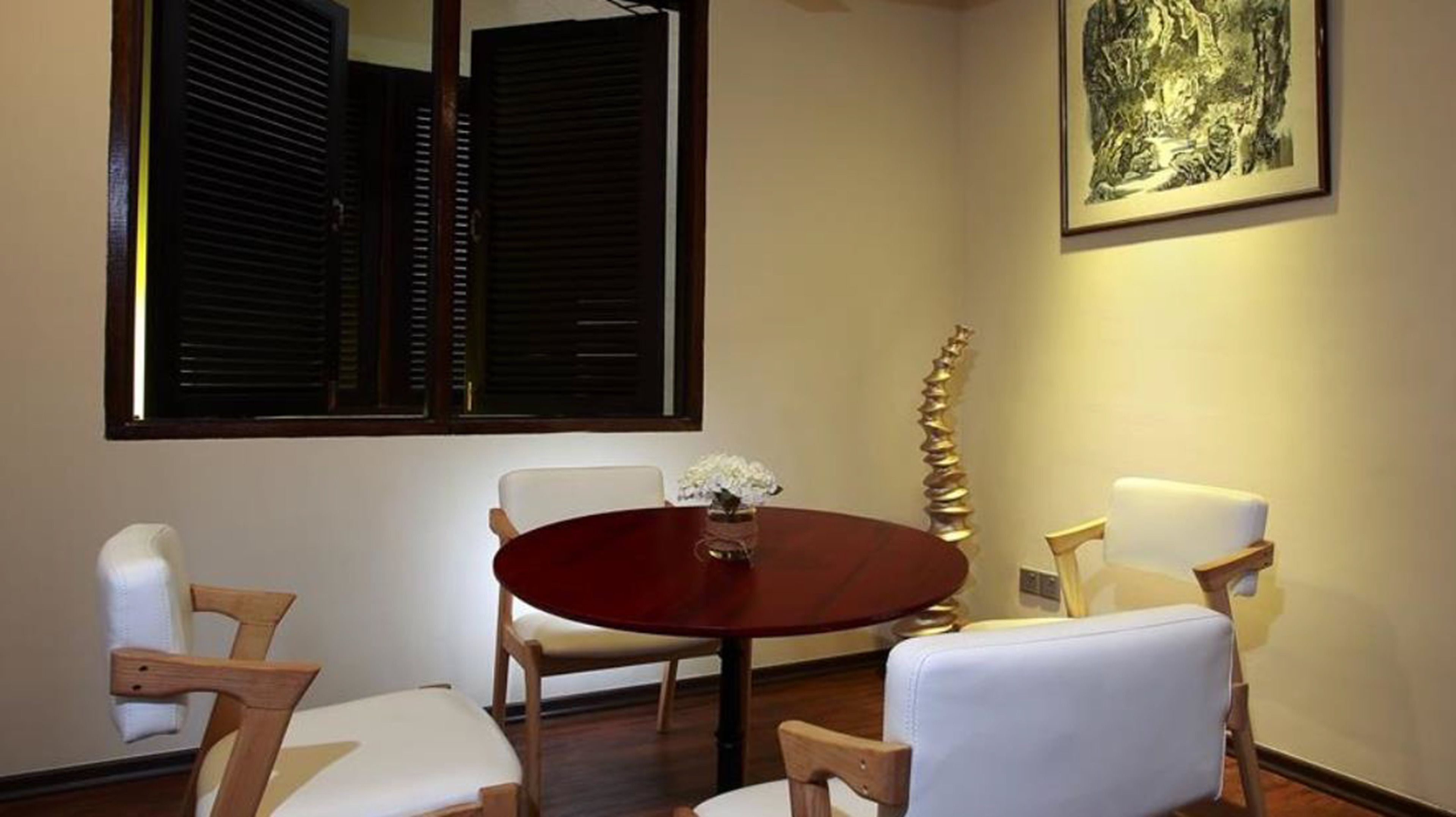 A dining area with a round table, four chairs, a vase with flowers, a window with shutters, and a framed artwork on the wall.