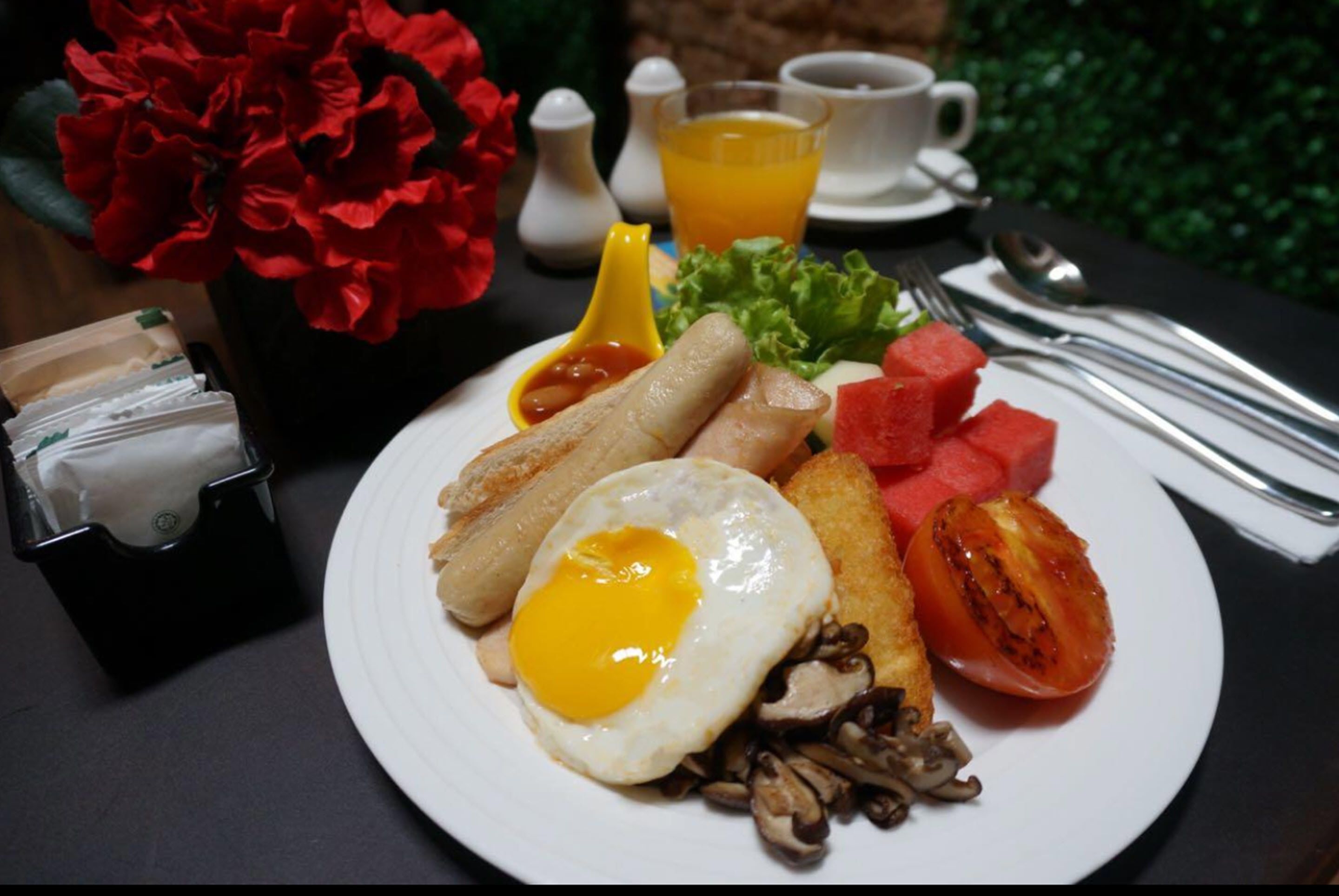 A breakfast plate with eggs, sausages, mushrooms, tomatoes, and watermelon, accompanied by a cup of orange juice and a vase of red flowers.