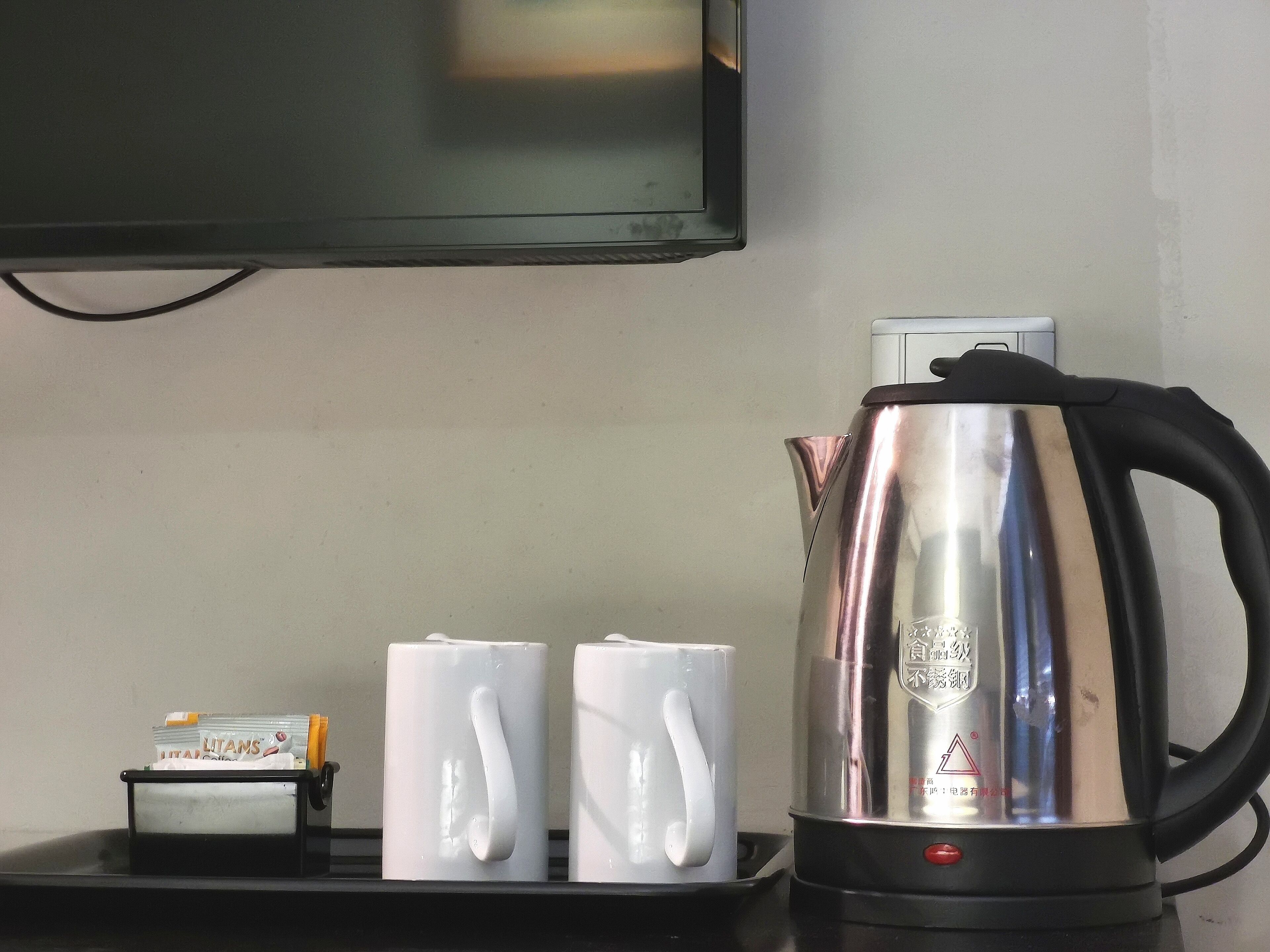 A stainless steel electric kettle on a black tray with two white mugs, a small box of cigarettes, and a television mounted on the wall.
