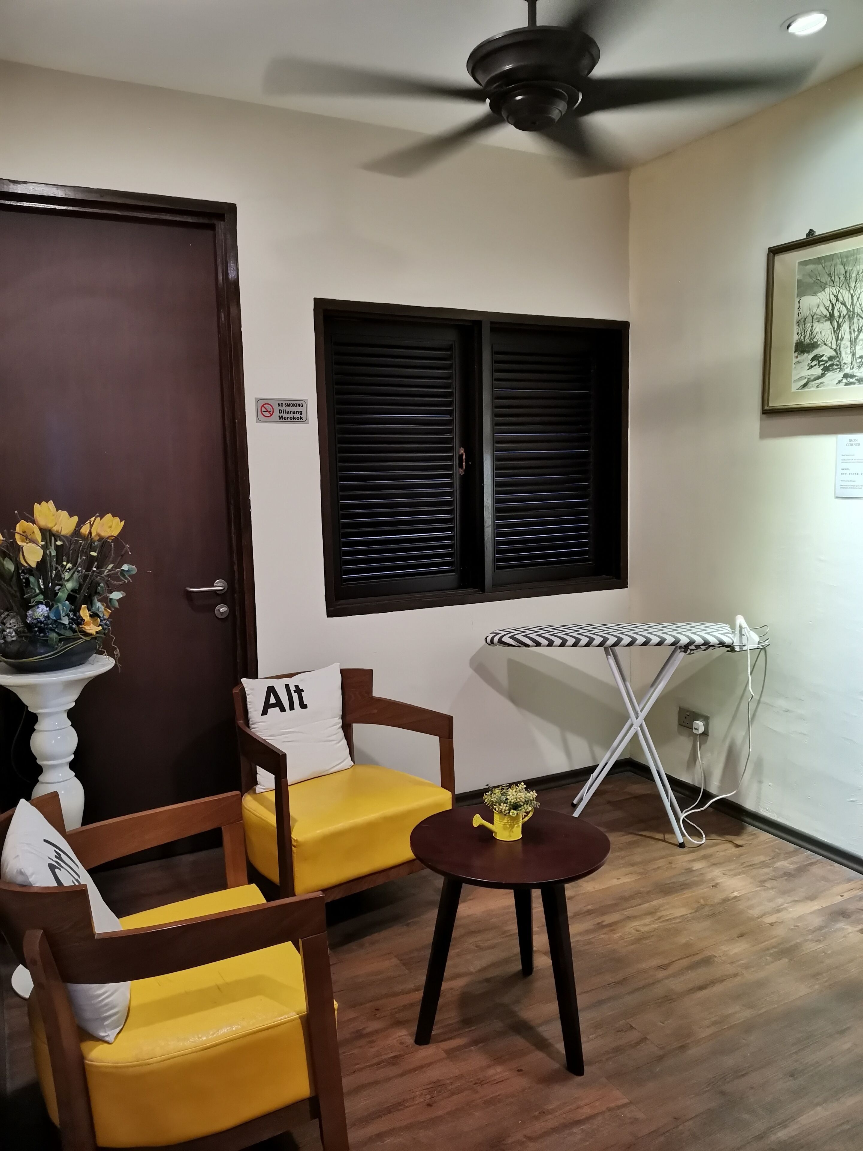 A room with a wooden door, a ceiling fan, a window with black shutters, a small round table with an ironing board, and a yellow sofa with a pillow labeled "Alt".