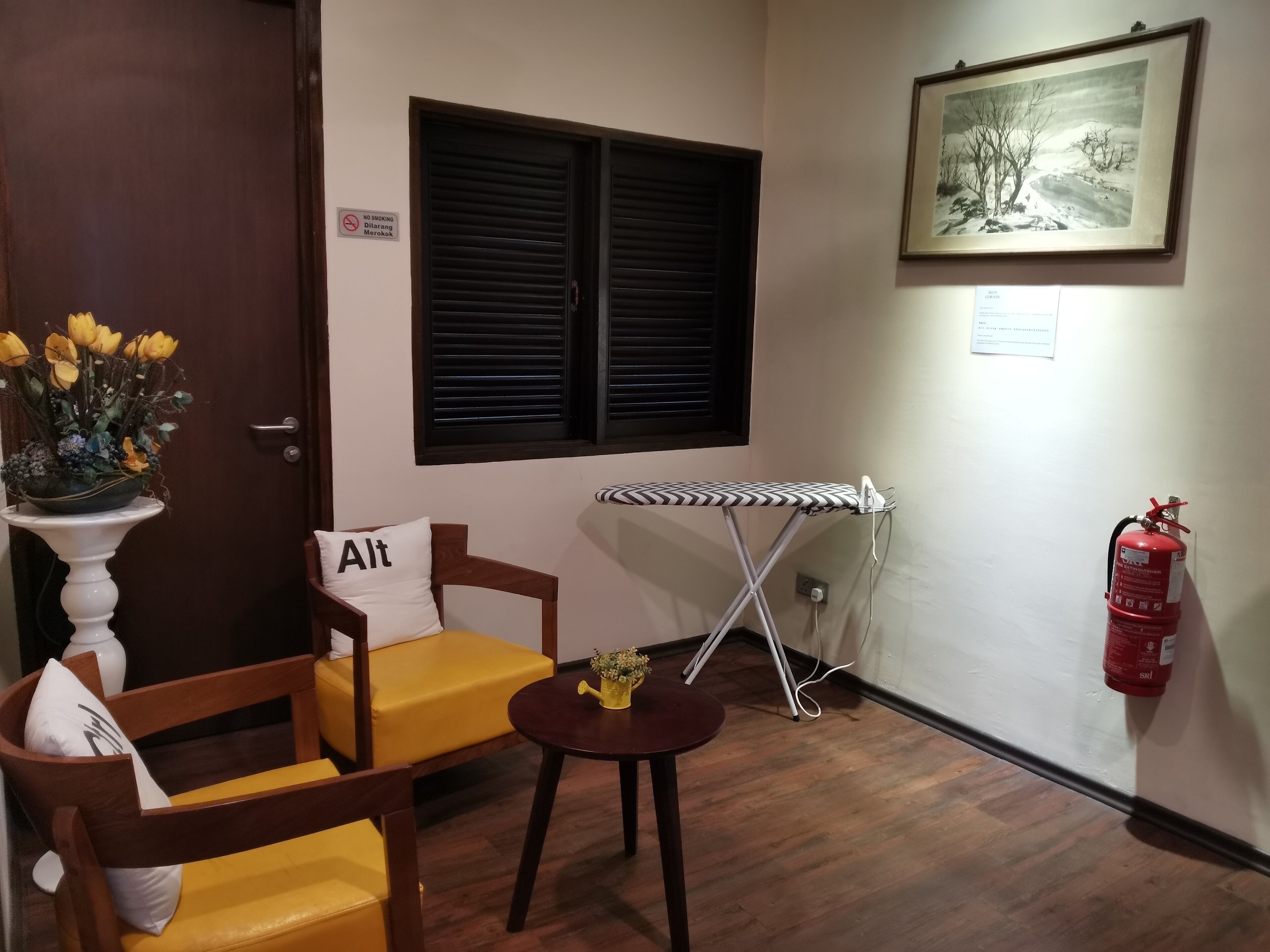 Una habitación con una silla de madera, una mesita redonda pequeña, una tabla de planchar fijada a la pared, un extintor y un cuadro enmarcado.