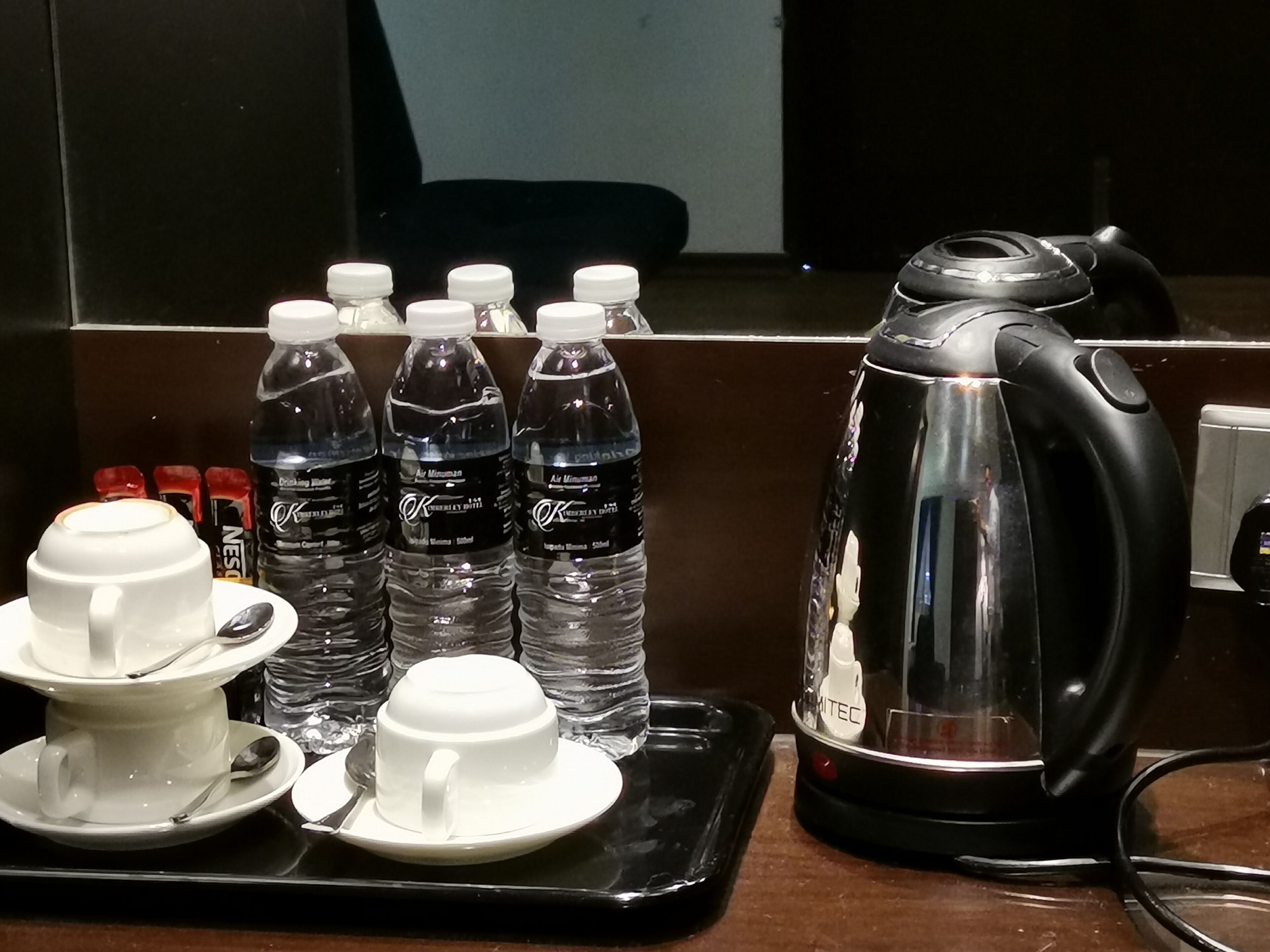 A tray with two white cups and saucers, a black electric kettle, and several water bottles with white caps.