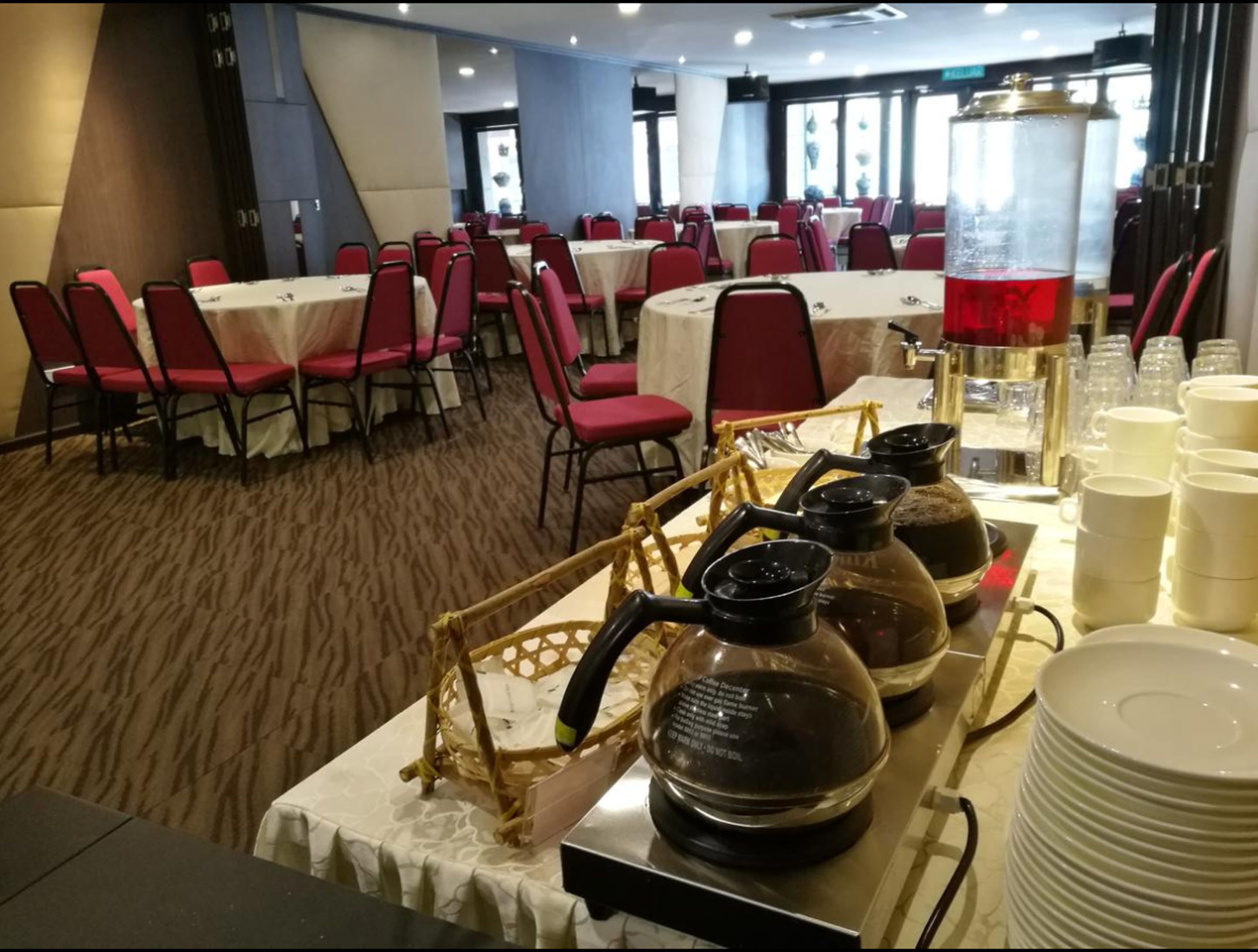 A banquet hall with tables set for dining, red chairs, and a coffee station with kettles and cups.