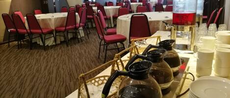 A banquet hall with tables set for dining, red chairs, and a coffee station with kettles and cups.