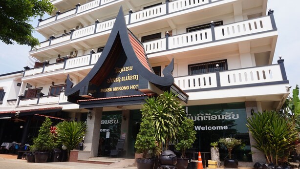 Un immeuble à plusieurs étages, de style architectural thaïlandais traditionnel, caractérisé par un toit noir et rouge bien visible, des balcons et des plantes en pot devant l’entrée.