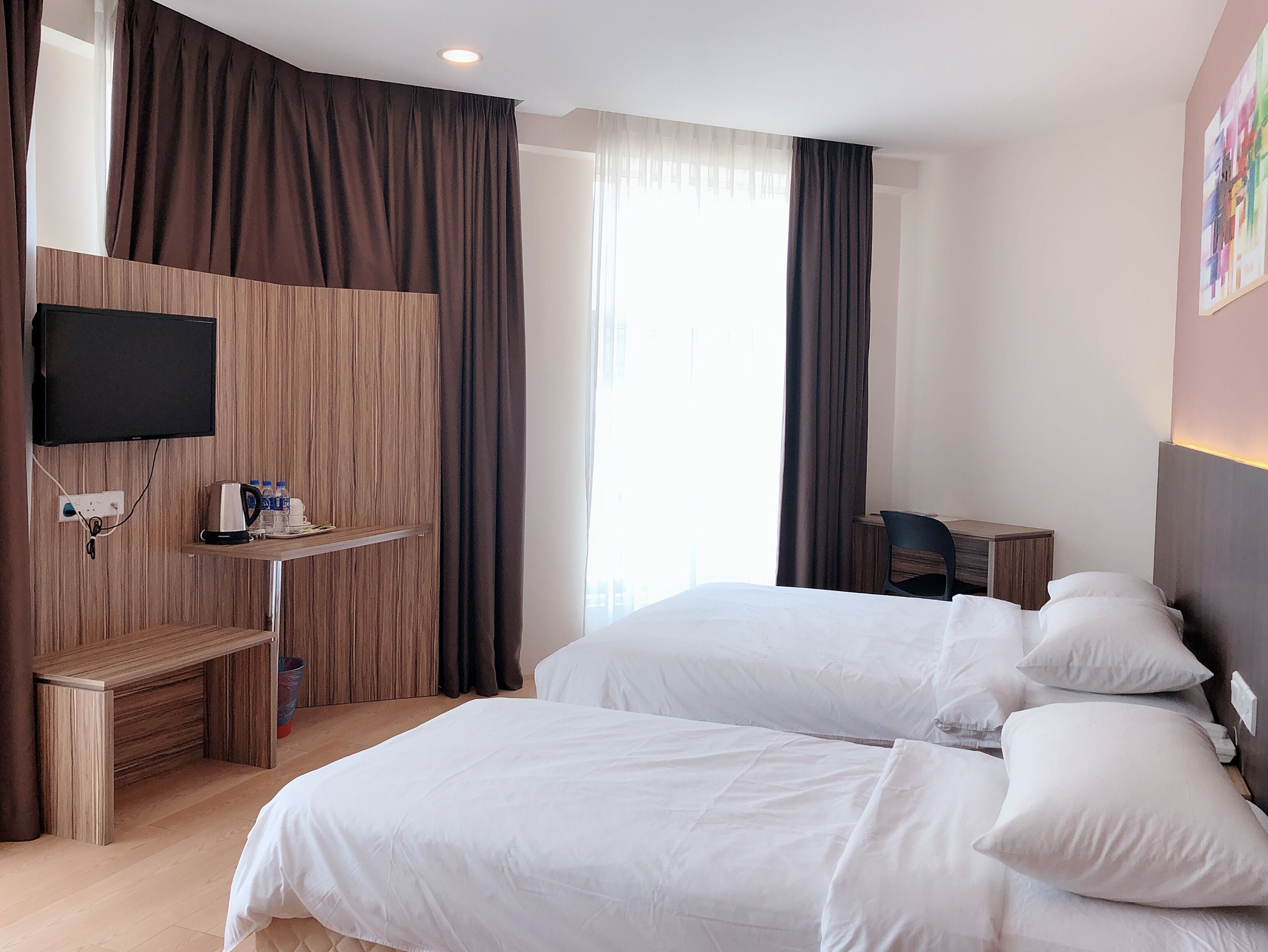 A hotel room with two beds, a TV mounted on the wall, a small desk with a chair, and a window with curtains.